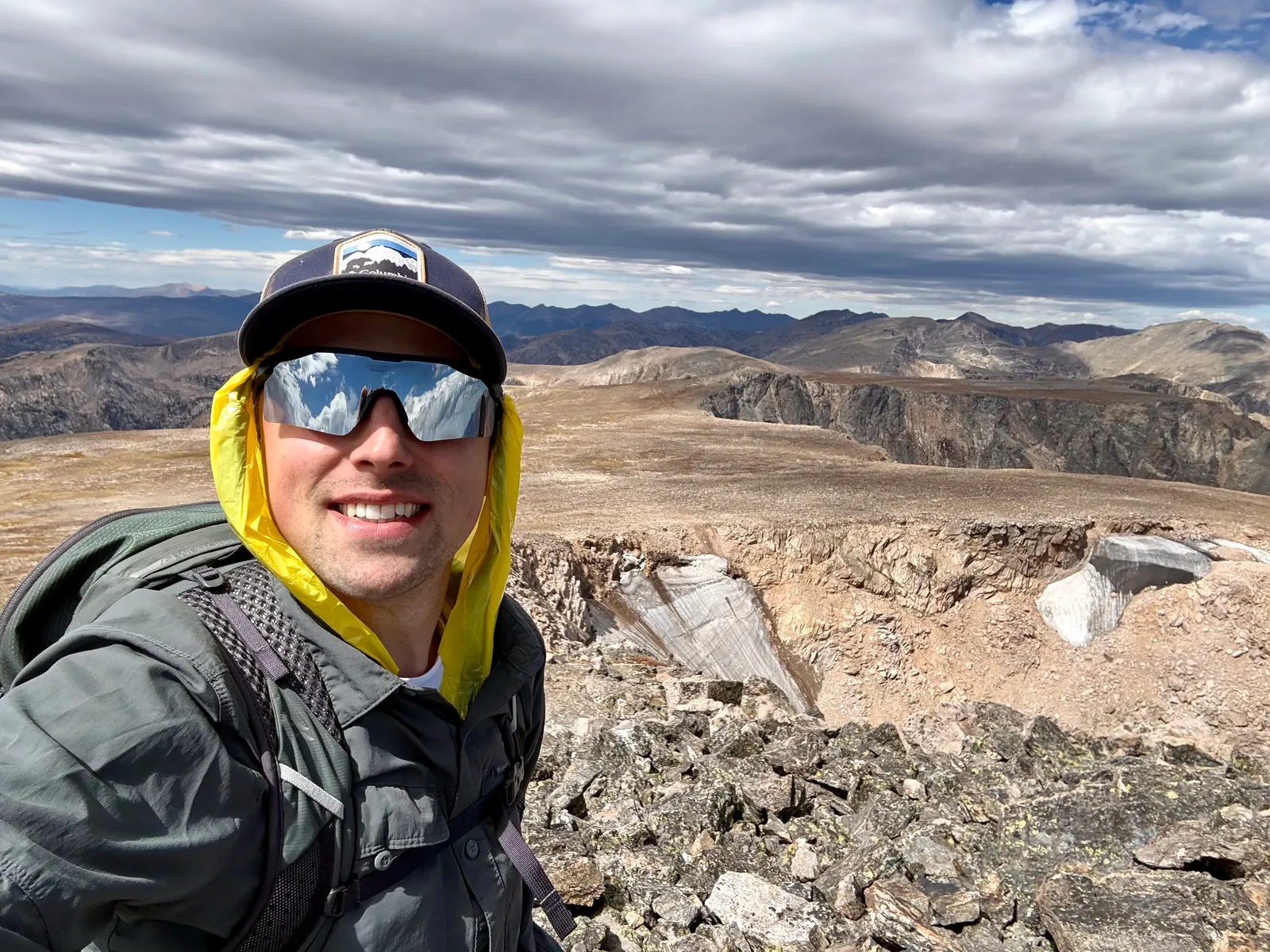 Me on the summit of the Little Matterhorn, smiling, in a cap, mirrored sunglasses and a yellow rain hood, with the rocky tundra and peaks of Rocky Mountain National Park behind me