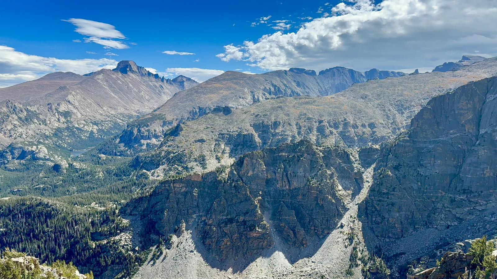 View from the Little Matterhorn: a deep forested valley with a small lake, grey rock walls in the foreground and Longs Peak on the skyline under a blue sky